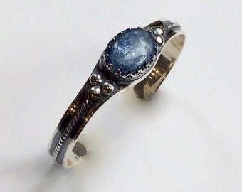 May include: A silver cuff bracelet with a blue gemstone set in a silver bezel. The bracelet has a textured design and small silver beads.