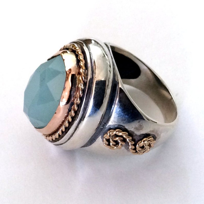 May include: A silver ring with a large, faceted, light blue gemstone set in a gold bezel. The ring band is decorated with a gold rope design.