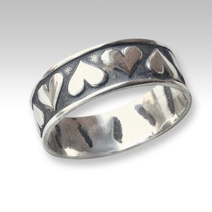 May include: A silver band ring with a textured finish and a row of raised heart designs around the band.