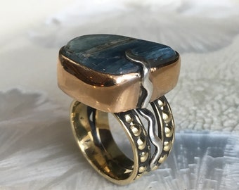 Puede incluir: Un anillo con una gran piedra preciosa azul verdosa engastada en un bisel de cobre. La banda está hecha de latón y plata, con un diseño texturizado.