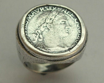 May include: A silver ring with a coin-shaped design featuring a profile of a woman's face and the inscription "MAVRSEXALE".