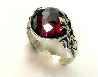 May include: A silver ring with a large, red gemstone set in a decorative, ornate setting. The ring features a fleur-de-lis design and small gold accents.