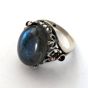 May include: A silver ring featuring a large oval blue labradorite gemstone set in a filigree design. The ring has small black and copper accents.