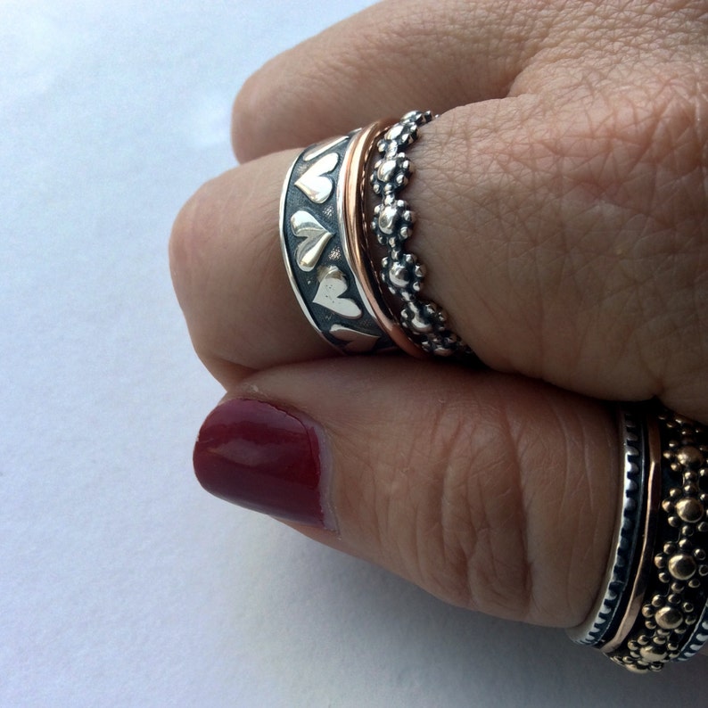 May include: A silver ring with a band of hearts, some of which are filled with gold. The ring is worn on a finger with two other rings, one silver and one gold.