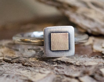 May include: A silver ring with a square setting. The setting has a silver border with a gold square in the center. The ring is on a wooden surface.