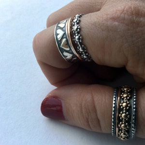 May include: Two silver rings with a copper band in the center. The rings are decorated with small hearts and dots. The rings are worn on a person's finger.