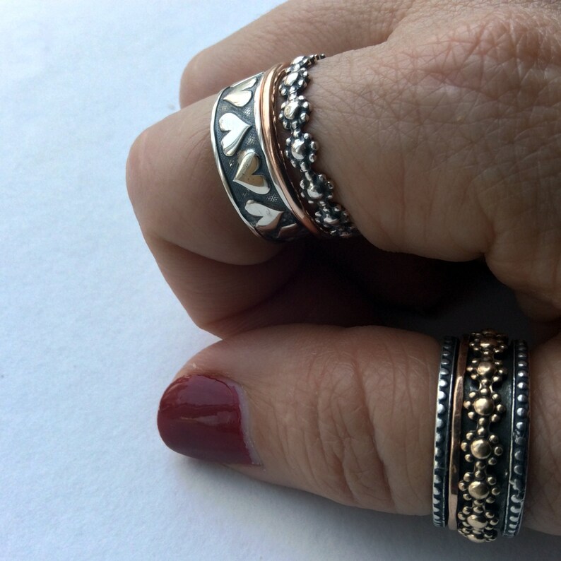 May include: Two silver rings with a copper band in the center. The rings are decorated with small hearts and dots. The rings are worn on a person's finger.