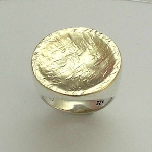 Può includere: Un anello d'argento con una parte superiore rotonda e testurizzata. La parte superiore dell'anello è un design martellato e testurizzato. L'anello è contrassegnato "925" sul lato.