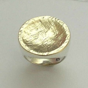 Può includere: Un anello d'argento con una parte superiore rotonda e testurizzata. L'anello è contrassegnato dal numero "925" sul lato.