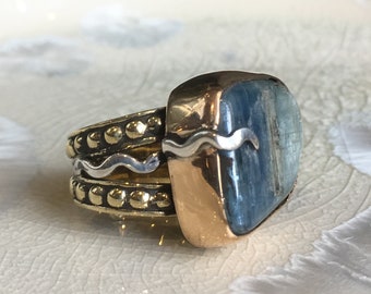 Puede incluir: Un conjunto de anillos, uno con una gran piedra azul rectangular con detalles dorados y plateados. Los otros anillos tienen cuentas doradas y diseños ondulados plateados. Los anillos se muestran sobre una superficie blanca texturizada.