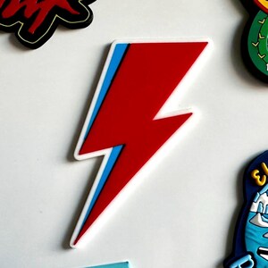 Puede incluir: Una pegatina roja en forma de rayo con un contorno azul y blanco. La pegatina es un diseño vibrante y llamativo, perfecto para decorar varios artículos. El diseño es una representación gráfica audaz de un rayo.