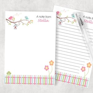 May include: A set of two blank note cards with a whimsical bird and flower design. The cards feature a pink bird, blue bird, and a birdhouse on a branch with green leaves and pink flowers. The bottom of the cards feature a green fence with pink, blue, and green pickets. The text "A note from Stella" is printed on both cards.