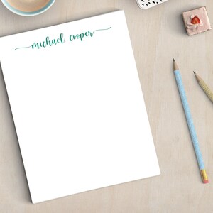 May include: A white notepad with the name "michael cooper" written in teal cursive at the top. The notepad is on a light wood surface with two pencils, one blue and one gold, and a small pink cake with a strawberry on top.