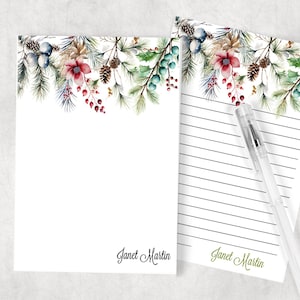 May include: A set of two personalized stationery notepads with a watercolor design of pine branches, berries, and flowers. The notepads have a white background with green, red, and blue accents. The text "Janet Martin" is printed at the bottom of each notepad.