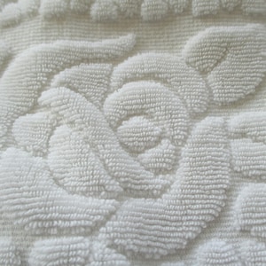 May include: Close-up of a white, textured towel with a raised floral pattern. The design features detailed, three-dimensional roses and leaves, creating a soft, plush appearance. The towel's surface is densely woven, providing a luxurious feel.