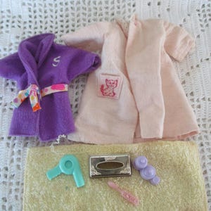 May include: A miniature spa set featuring a purple robe with a white "S" and a patterned belt, a pink robe with a cat patch, a yellow towel, a teal hairdryer, a silver compact, a pink brush, and purple rollers. The set is arranged on a white crocheted surface.