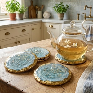May include: A clear glass teapot filled with tea and white flowers sits on a round blue coaster with gold-leaf detailing and a textured gold rim. Three additional matching blue and gold coasters are arranged nearby on the wooden surface.