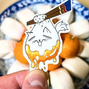 以下が含まれることがあります： 白い漫画の猫の形をしたステッカーで、頭にはゴマの入ったパンが乗っています。猫は黄色のソースを垂らしており、ステッカーは白いパンの皿の上に置かれています。