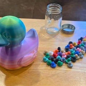 May include: A large, colorful 3D-printed duck with a gradient of blue, green, and purple hues. A clear glass jar with a metal lid and a collection of small, colorful duck-shaped objects are also present on a wooden surface.