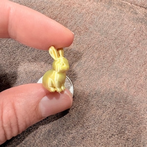 May include: A small, gold-colored 3D-printed bunny figurine. The bunny is sitting on a coin and held between two fingers. The figurine has detailed features, including ears, eyes, and paws. The background is a brown, textured surface.