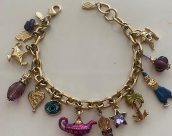 May include: Gold-tone charm bracelet featuring various charms, including a genie lamp, a palm tree, a star, and an evil eye. The bracelet has a lobster clasp closure and is adorned with purple, blue, and gold accents. The charms are intricately detailed.