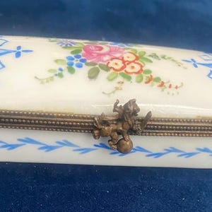 May include: A white porcelain box with a blue and white floral design and a gold trim. The box has a hinged lid with a brass lion finial.