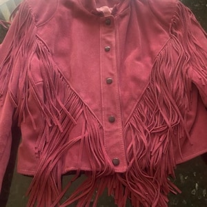 May include: A dusty rose suede jacket with fringe detailing. The jacket features a round collar, button closure, and long fringe along the front and sleeves. The jacket is a western-inspired style.