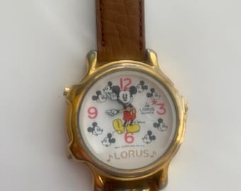 May include: A gold-tone Mickey Mouse watch with a white face and brown leather band. The watch features Mickey Mouse and other Mickey Mouse characters around the face. The watch face also says "Lorus Quartz" and "Disney".