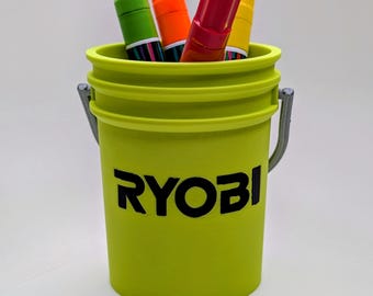 May include: A lime green bucket with the word "RYOBI" in black lettering. The bucket contains four markers in green, orange, red, and yellow. The bucket has a metal handle.
