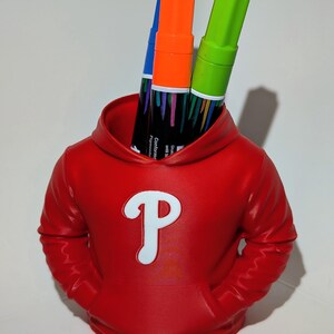 May include: A red pen holder shaped like a hoodie, featuring a white "P" logo. The holder contains three markers: blue, orange, and green. The markers have black labels with text. The pen holder has two pockets.