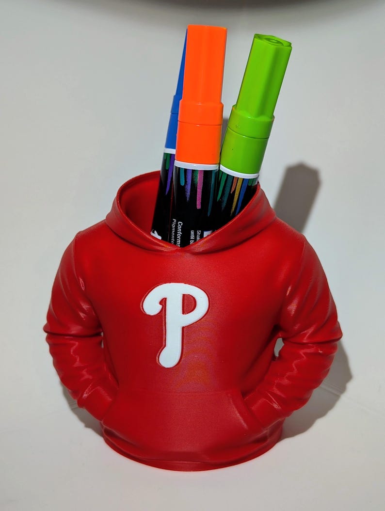 May include: A red pen holder shaped like a hoodie, featuring a white "P" logo. The holder contains three markers: blue, orange, and green. The markers have black labels with text. The pen holder has two pockets.