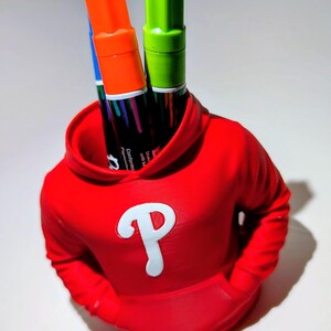 May include: A red hoodie-shaped pen holder with a white "P" logo, holding orange and green markers. The pen holder has a pocket design and is on a white surface.