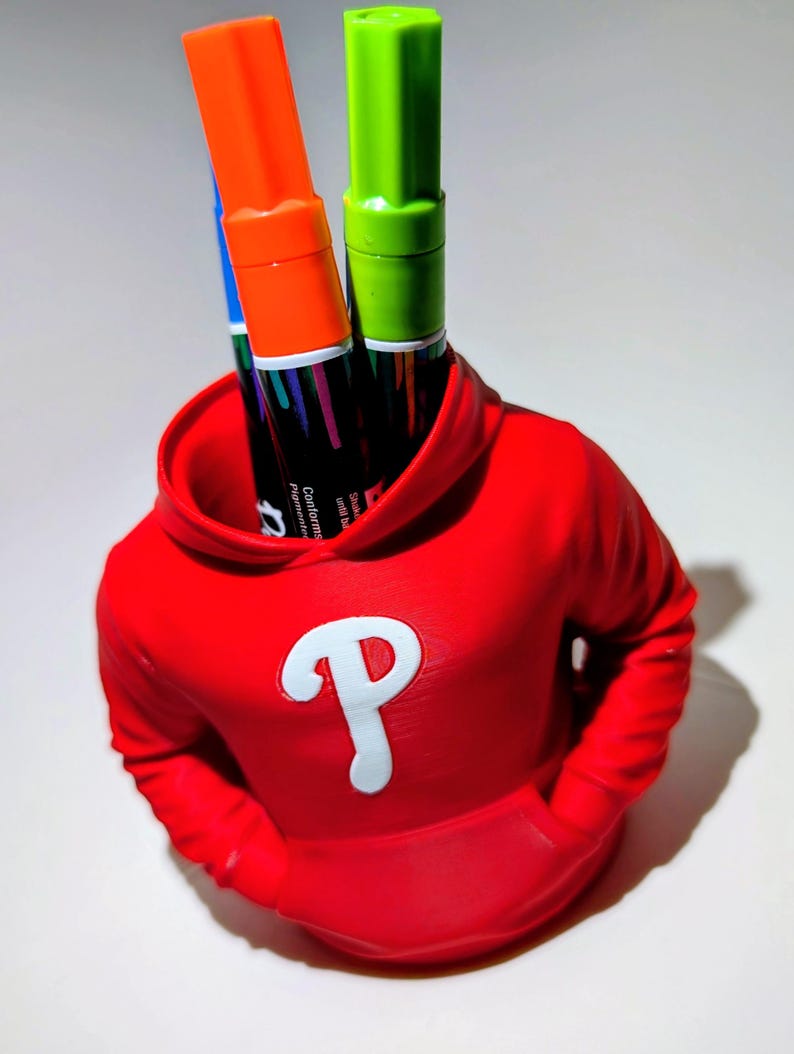 May include: A red hoodie-shaped pen holder with a white "P" logo, holding orange and green markers. The pen holder has a pocket design and is on a white surface.