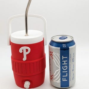 May include: A red and white plastic beverage cooler with a white handle, a white lid, a white spigot, and a metal straw inserted through the top. Next to it stands a silver and blue 12 fl oz aluminum can labeled Flight Next Generation of Light Beer.