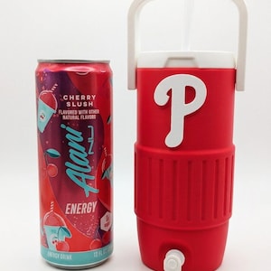 May include: A 12 fluid ounce Alani Nu Cherry Slush energy drink can next to a red insulated beverage cooler with a white handle, a white spigot at the base, and a large white letter P emblem on the front, featuring a clear straw extending from the top.