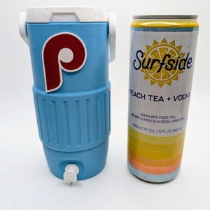 May include: A light blue beverage cooler with a white lid, handle, and spigot, featuring a red and white P logo. Next to it is a 12 fl. oz. Surfside Peach Tea + Vodka can with a yellow sun graphic, white background, and horizontal color stripes.