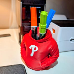 May include: A red pen holder shaped like a hoodie, with a white "P" logo on the front. The holder contains an orange, green, and blue marker. The pen holder is on a white surface.