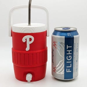May include: A red and white plastic beverage cooler with a white handle, a white lid, and a white spigot, featuring a white letter P logo on the front. A silver metal straw protrudes from the top. Next to it is a 12 fluid ounce aluminum beer can.