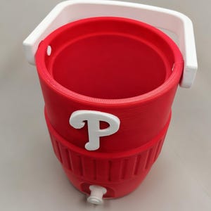 May include: A red cylindrical beverage cooler featuring a white handle and a white spigot at the base. The exterior displays a prominent white letter P emblem on the front. The cooler has a ribbed texture around the lower section and an open top.
