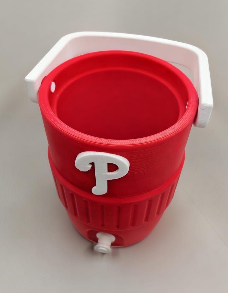 May include: A red cylindrical beverage cooler featuring a white handle and a white spigot at the base. The exterior displays a prominent white letter P emblem on the front. The cooler has a ribbed texture around the lower section and an open top.