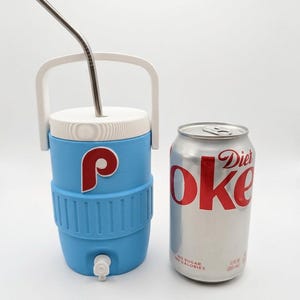 May include: A light blue beverage cooler with a white handle, white lid, and a white spigot, featuring a red and white letter P logo. A silver metal straw is inserted into the lid. Next to it is a 12 fluid ounce silver Diet Coke aluminum can.