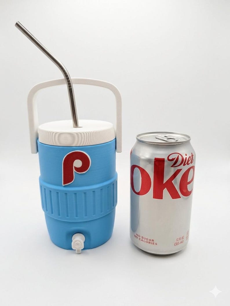 May include: A light blue beverage cooler with a white handle, white lid, and a white spigot, featuring a red and white letter P logo. A silver metal straw is inserted into the lid. Next to it is a 12 fluid ounce silver Diet Coke aluminum can.