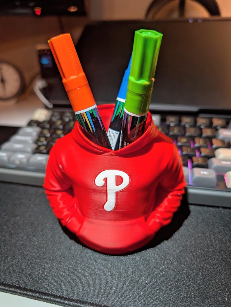 May include: A red pen holder shaped like a hoodie, with a white "P" logo on the front. The holder contains an orange, green, and blue marker. The pen holder is on a black surface, with a keyboard in the background.