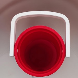 May include: A top-down view of a cylindrical red plastic bucket featuring a white handle attached to the sides. The interior of the bucket is hollow and red, while the exterior rim and the arched handle are white, creating a high-contrast design.