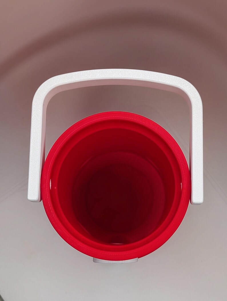 May include: A top-down view of a cylindrical red plastic bucket featuring a white handle attached to the sides. The interior of the bucket is hollow and red, while the exterior rim and the arched handle are white, creating a high-contrast design.