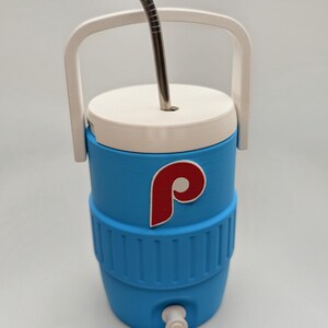 May include: A light blue beverage cooler with a white lid, a white handle, and a white spigot at the base. The cooler features a red and white letter P logo on the front and includes a silver metal straw inserted through the top lid.
