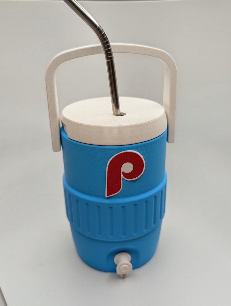 May include: A light blue beverage cooler with a white lid, a white handle, and a white spigot at the base. The cooler features a red and white letter P logo on the front and includes a silver metal straw inserted through the top lid.