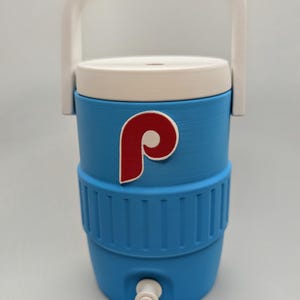 May include: A light blue beverage cooler with a white lid and a white handle. The cooler features a red and white letter P logo on the front and a white spigot at the base. The exterior has a ribbed texture around the middle section of the container.