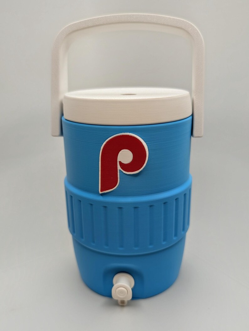 May include: A light blue beverage cooler with a white lid and a white handle. The cooler features a red and white letter P logo on the front and a white spigot at the base. The exterior has a ribbed texture around the middle section of the container.