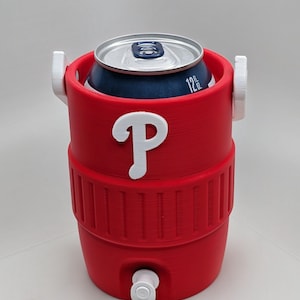 May include: A red cylindrical beverage cooler designed to hold a 12 fl oz can, featuring a white letter P logo on the front, two white side handles, and a small white spigot at the base. The cooler has a ribbed texture around the middle section.